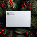 A white textured paper envelope or card centered in a holiday flat-lay composition, surrounded by green pine branches and red berries. The top-left corner shows a printed black logo and address reading 'The Spice' with a Christmas tree icon.