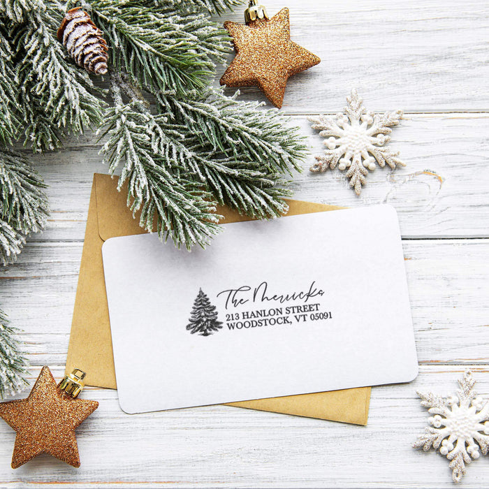 A white holiday-themed address card with black printed text and a small pine tree logo, placed on top of a kraft paper envelope. The card is styled in a flat lay with frosted green pine branches, glittery gold star ornaments, white snowflake ornaments, and a rustic white wood plank background.