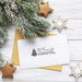 A white holiday-themed address card with black printed text and a small pine tree logo, placed on top of a kraft paper envelope. The card is styled in a flat lay with frosted green pine branches, glittery gold star ornaments, white snowflake ornaments, and a rustic white wood plank background.