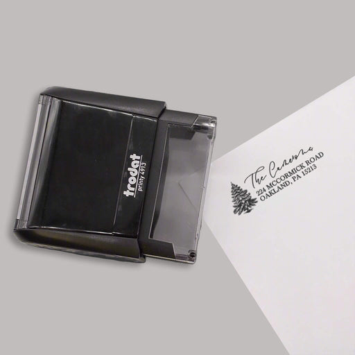 A black self-inking stamp shown on a gray background, with a sample stamped address printed on a white sheet of paper.