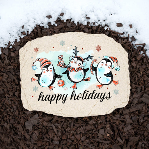 A decorative holiday garden stone with a penguin illustration and the words 'happy holidays' placed on dark wood mulch with snow at the top.