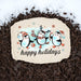 A decorative holiday garden stone with a penguin illustration and the words 'happy holidays' placed on dark wood mulch with snow at the top.