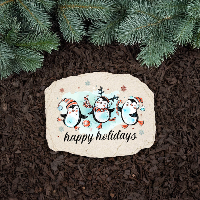 A decorative rectangular stone plaque with rounded corners sits on dark brown mulch beneath green pine branches. The plaque features an illustration of three penguins wearing colorful winter hats and scarves, with the words 'happy holidays' printed below in black script.