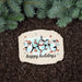 A decorative rectangular stone plaque with rounded corners sits on dark brown mulch beneath green pine branches. The plaque features an illustration of three penguins wearing colorful winter hats and scarves, with the words 'happy holidays' printed below in black script.