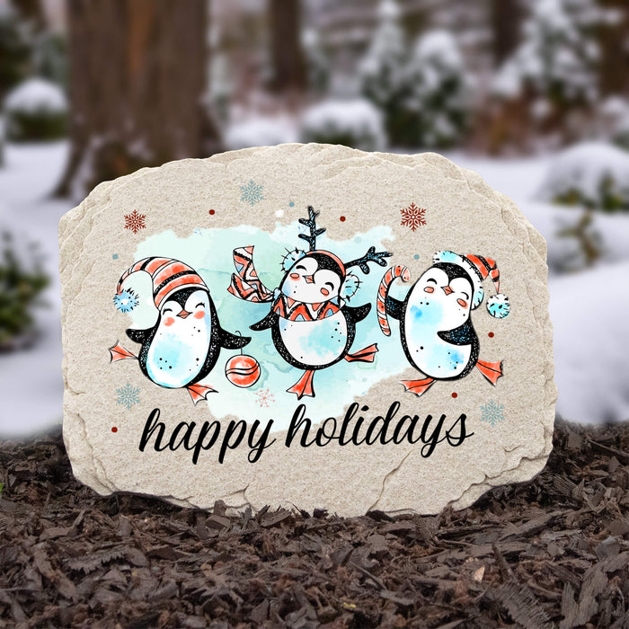 A decorative garden stone with a holiday penguin illustration and the words 'happy holidays' in black script, placed outdoors on mulch with a snowy blurred background.