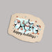 A decorative stone plaque with irregular edges, featuring an illustration of three penguins in winter hats and scarves on a light blue watercolor background, with the words 'happy holidays' in black script. The plaque is photographed on a plain light gray background.