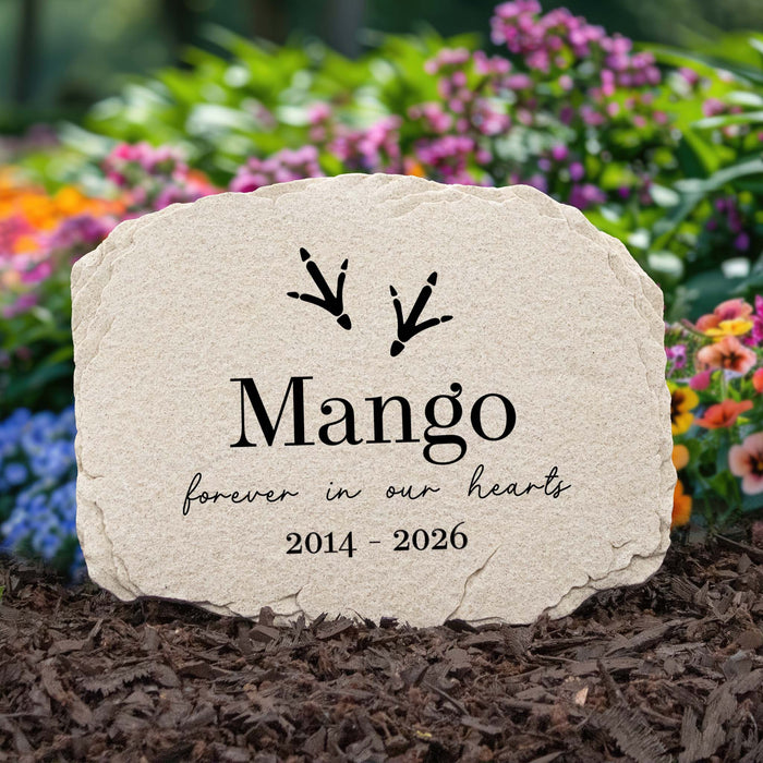 Personalized pet memorial garden stone with black engraved text reading 'Mango', 'forever in our hearts', and '2014 - 2026', plus two small bird footprint marks, shown standing in mulch with blurred flowers in the background.