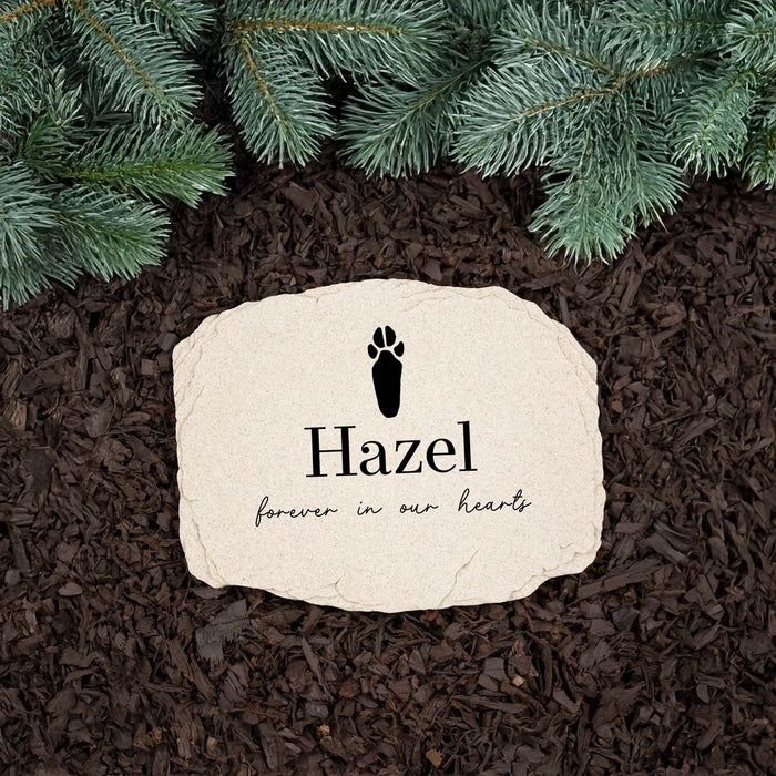A personalized pet bunny memorial garden stone with a black rabbit paw print and the name 'Hazel' on a natural beige stone, placed on dark brown mulch with green pine branches at the top.
