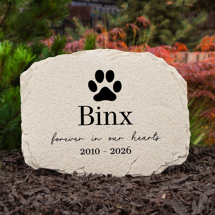 A personalized pet memorial stone with a black cat paw print and the name 'Binx' engraved, surrounded by mulch and greenery in an outdoor setting.