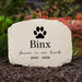 A personalized pet memorial stone with a black cat paw print and the name 'Binx' engraved, surrounded by mulch and greenery in an outdoor setting.