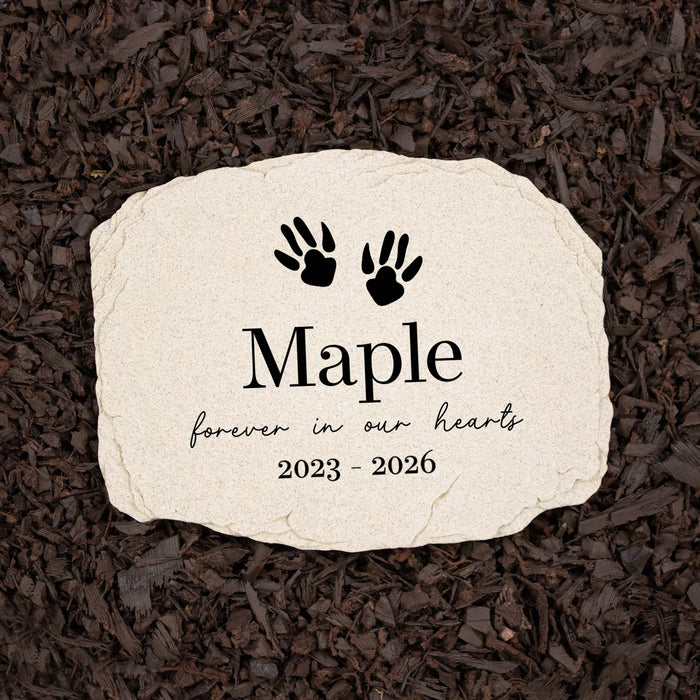 Personalized pet memorial garden stone with black hamster paw prints, the name 'Maple', and the years '2023 - 2026' on a light beige stone, placed on a bed of dark brown mulch.