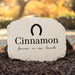 A personalized light beige stone memorial garden marker with a black horseshoe graphic and the name 'Cinnamon' along with the phrase 'forever in our hearts', displayed outdoors on dark mulch with a blurred autumn garden background.