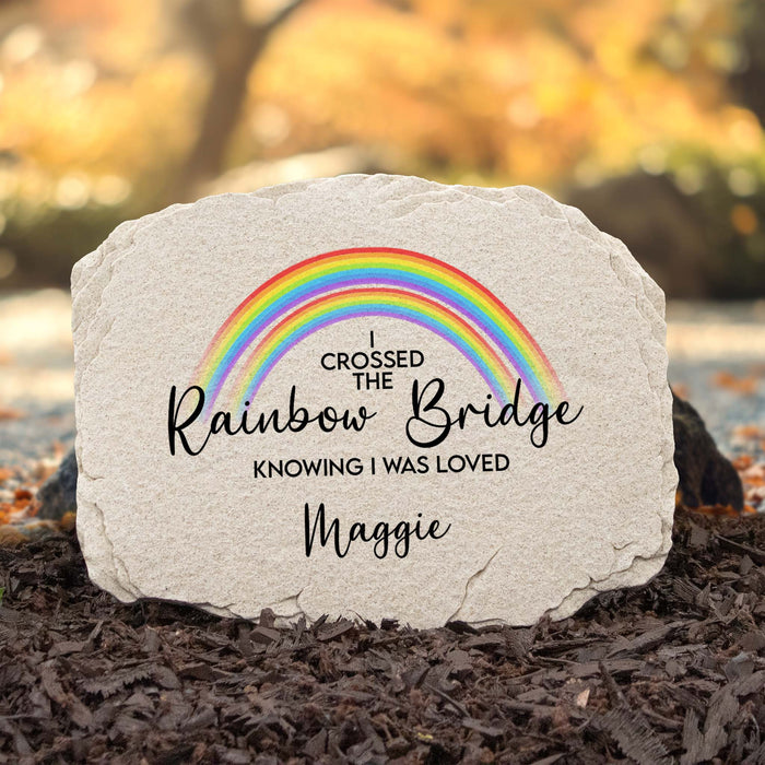 Personalized memorial garden stone with a printed rainbow and the text 'I Crossed the Rainbow Bridge Knowing I Was Loved Maggie' set in mulch outdoors.