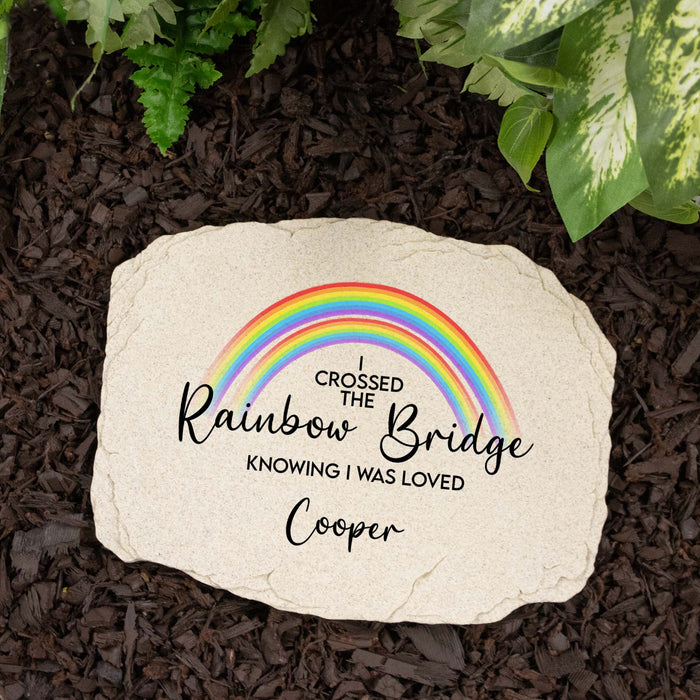 A personalized garden stone with a printed rainbow and the text 'I Crossed the Rainbow Bridge Knowing I Was Loved Cooper' placed on dark brown mulch with green plants around it.