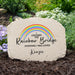 Personalized beige memorial garden stone with a printed rainbow and the text 'I Crossed the Rainbow Bridge Knowing I Was Loved' and the name 'Kenzie,' shown outdoors in mulch with greenery and flowers in the background.