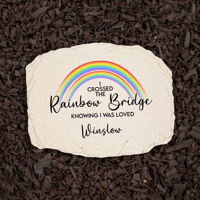 A personalized cream-colored stone garden plaque with a colorful rainbow graphic and the text 'I CROSSED THE Rainbow Bridge KNOWING I WAS LOVED Winslow', placed on a bed of dark brown mulch.