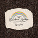 A personalized cream-colored stone garden plaque with a colorful rainbow graphic and the text 'I CROSSED THE Rainbow Bridge KNOWING I WAS LOVED Winslow', placed on a bed of dark brown mulch.