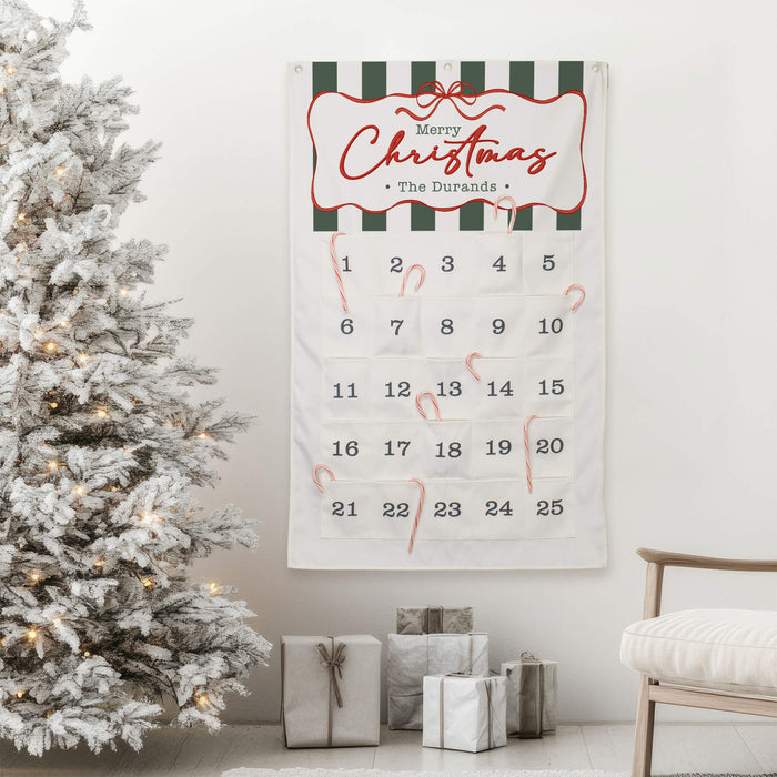 A Christmas advent calendar hanging on a white wall in a decorated living room, next to a flocked Christmas tree and wrapped gifts. The calendar is off-white with dark green and white stripes at the top, red cursive 'Merry Christmas' lettering, numbered pockets, and several red candy canes.