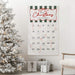 A Christmas advent calendar hanging on a white wall in a decorated living room, next to a flocked Christmas tree and wrapped gifts. The calendar is off-white with dark green and white stripes at the top, red cursive 'Merry Christmas' lettering, numbered pockets, and several red candy canes.