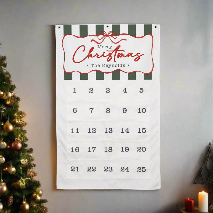 A personalized Christmas advent calendar hanging on a gray wall, flanked by a decorated Christmas tree on the left and candles on the right. The white fabric calendar features dark green and white stripes at the top, a red decorative border with 'Merry Christmas The Reynolds' written in red script, and 25 numbered pockets for the days.
