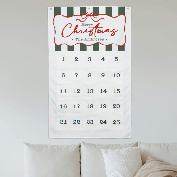 A personalized Christmas advent calendar hanging on a light gray wall above a bed with white pillows. The tapestry features a white background with dark green and white horizontal stripes at the top, a red decorative border, and 'Merry Christmas - The Ambroses' written in red script.