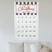 A personalized Christmas advent calendar hanging on a light gray wall above a bed with white pillows. The tapestry features a white background with dark green and white horizontal stripes at the top, a red decorative border, and 'Merry Christmas - The Ambroses' written in red script.