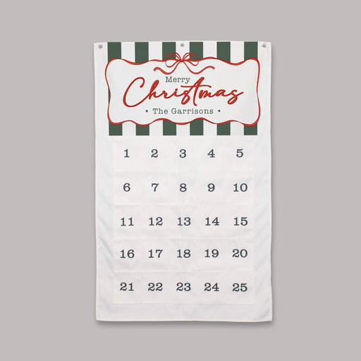 A personalized Christmas advent calendar featuring a white fabric body with dark green and white striped accents at the top, a red decorative border around the 'Merry Christmas The Garrisons' greeting, and 25 numbered pockets for the days of the week on a solid gray background.