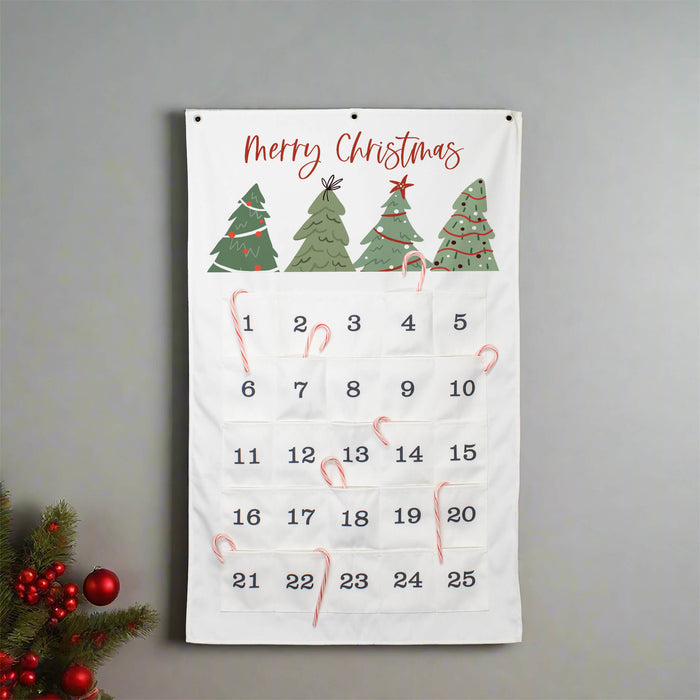 A white Christmas advent calendar hanging on a gray wall, with four illustrated green Christmas trees at the top, the words 'Merry Christmas' in red script, and 25 numbered pockets with red candy canes. A small decorated evergreen branch with red berries appears in the lower left corner.