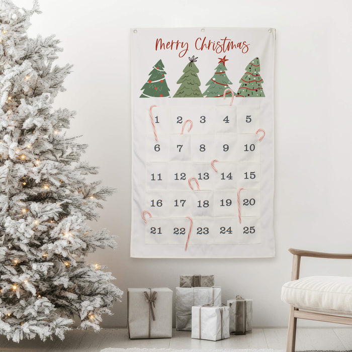 A Christmas advent calendar hanging on a white wall in a decorated living room, next to a flocked Christmas tree, wrapped gifts, and a chair. The calendar is white with green Christmas trees, red 'Merry Christmas' script, numbered pockets, and has candy canes in the pockets.