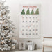 A Christmas advent calendar hanging on a white wall in a decorated living room, next to a flocked Christmas tree, wrapped gifts, and a chair. The calendar is white with green Christmas trees, red 'Merry Christmas' script, numbered pockets, and has candy canes in the pockets.