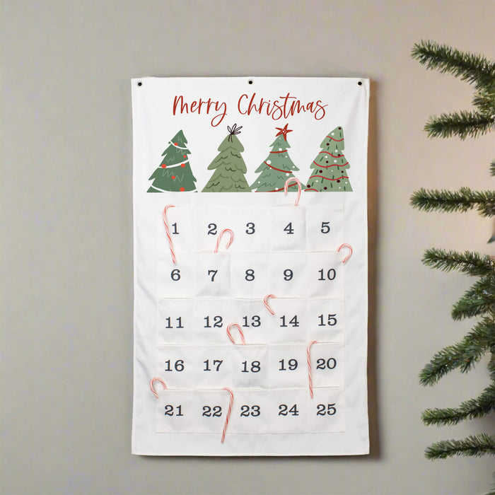 A white Christmas advent calendar hanging on a light gray wall, decorated with green Christmas trees. Pine tree branches are visible on the right side of the frame.