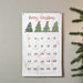 A white Christmas advent calendar hanging on a light gray wall, decorated with green Christmas trees. Pine tree branches are visible on the right side of the frame.