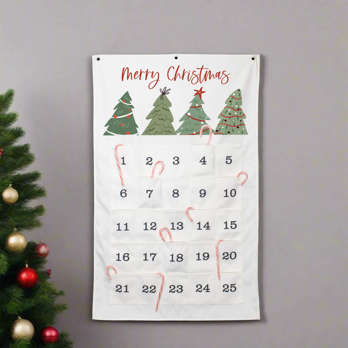 A white Christmas advent calendar with 'Merry Christmas' written at the top, decorated with four green Christmas trees and 25 numbered pockets. The calendar is hanging on a gray wall next to a decorated Christmas tree with red and gold ornaments.