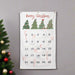 A white Christmas advent calendar with 'Merry Christmas' written at the top, decorated with four green Christmas trees and 25 numbered pockets. The calendar is hanging on a gray wall next to a decorated Christmas tree with red and gold ornaments.