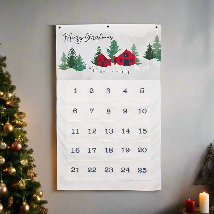 A personalized Christmas advent calendar hanging on a gray wall, decorated with a winter scene illustration of red cabins, pine trees, and the text 'Merry Christmas' and 'Jenkins Family'. The calendar shows 25 numbered pockets for advent days.