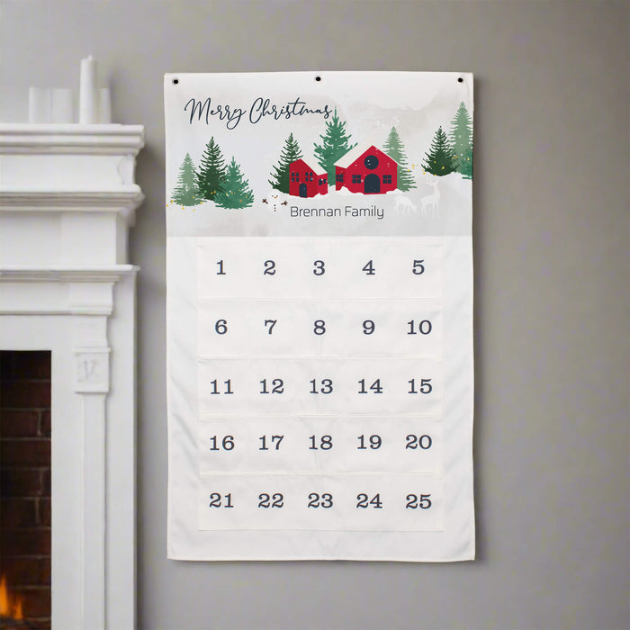 A personalized Christmas calendar hanging on a gray wall next to a white fireplace mantel. The white fabric calendar features a festive winter illustration at the top with the text 'Merry Christmas' and 'Brennan Family', followed by a grid of numbers from 1 to 25.