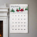 A personalized Christmas calendar hanging on a gray wall next to a white fireplace mantel. The white fabric calendar features a festive winter illustration at the top with the text 'Merry Christmas' and 'Brennan Family', followed by a grid of numbers from 1 to 25.