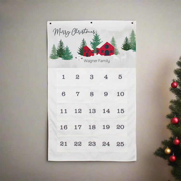 A personalized Christmas calendar hanging on a neutral gray wall, featuring a festive winter scene with pine trees, a red cabin, and the text 'Merry Christmas' and 'Wagner Family' above a numbered grid from 1 to 25. A decorated Christmas tree is partially visible on the right side.