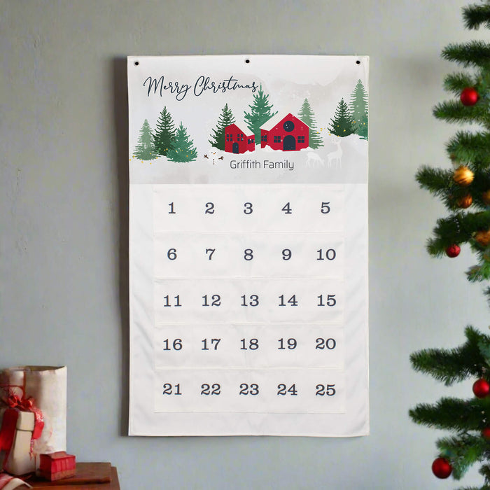 A personalized Christmas calendar hanging on a light gray wall, featuring a winter landscape illustration with a red cabin, evergreen trees, and the text 'Merry Christmas' and 'Griffith Family'. The calendar numbers are arranged in a grid on a white background.