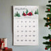 A personalized Christmas calendar hanging on a light gray wall, featuring a winter landscape illustration with a red cabin, evergreen trees, and the text 'Merry Christmas' and 'Griffith Family'. The calendar numbers are arranged in a grid on a white background.