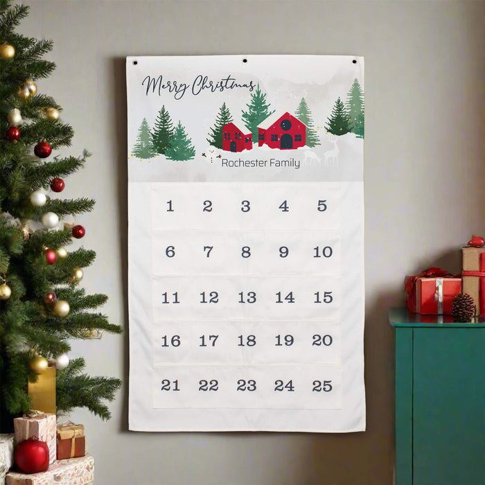 A personalized Christmas calendar hanging on a wall, featuring a festive winter scene with red cabins, pine trees, and the text 'Merry Christmas' and 'Rochester Family' above a numbered grid from 1 to 25. The calendar is styled in a holiday home setting next to a decorated Christmas tree and a teal cabinet with wrapped gifts.