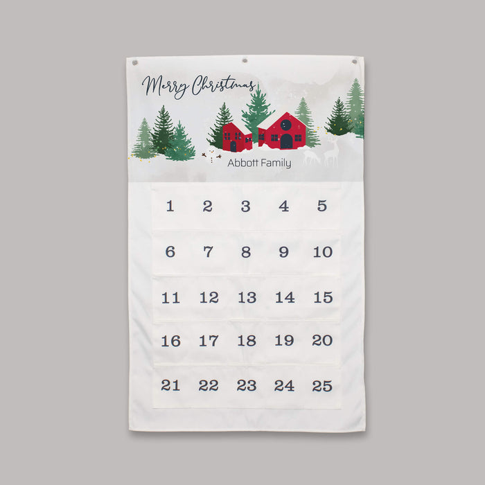 A personalized Christmas countdown calendar hanging on a neutral gray wall. The white fabric calendar features a festive winter illustration at the top with the text 'Merry Christmas' and 'Abbott Family' beneath a red cabin and pine trees.