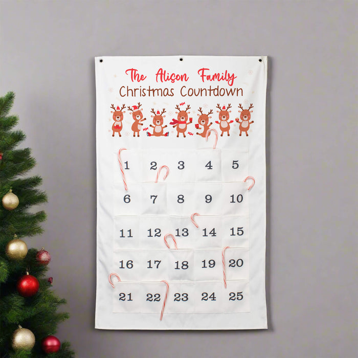 A personalized Christmas countdown calendar hanging on a gray wall next to a decorated Christmas tree. The white fabric calendar features the text 'The Alison Family Christmas Countdown' in red, a row of cartoon reindeer, and a numbered grid from 1 to 25 with candy canes tucked into some pockets.