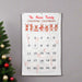 A personalized Christmas countdown calendar hanging on a gray wall next to a decorated Christmas tree. The white fabric calendar features the text 'The Alison Family Christmas Countdown' in red, a row of cartoon reindeer, and a numbered grid from 1 to 25 with candy canes tucked into some pockets.