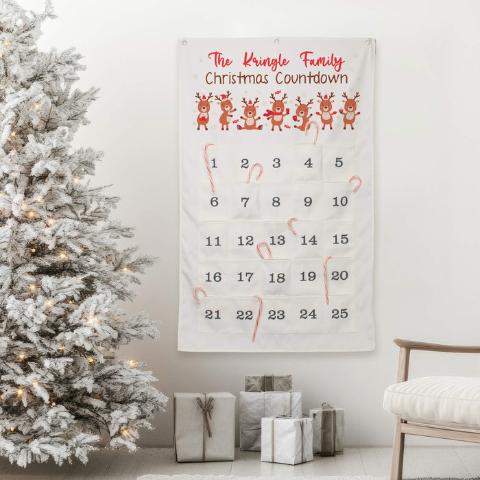 A personalized Christmas advent calendar hanging on a wall in a decorated living room, next to a flocked Christmas tree and wrapped gifts. The white fabric calendar features red text reading 'The Kingle Family Christmas Countdown' at the top, a row of illustrated reindeer, and numbered pockets from 1 to 25 with candy canes in some of the pockets.