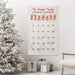 A personalized Christmas advent calendar hanging on a wall in a decorated living room, next to a flocked Christmas tree and wrapped gifts. The white fabric calendar features red text reading 'The Kingle Family Christmas Countdown' at the top, a row of illustrated reindeer, and numbered pockets from 1 to 25 with candy canes in some of the pockets.