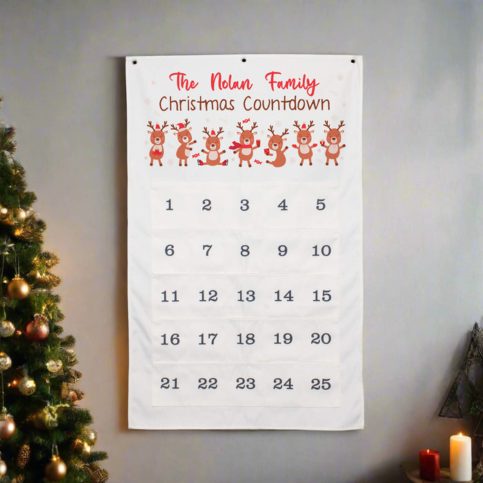 A personalized Christmas countdown calendar hanging on a gray wall next to a decorated Christmas tree. The white fabric calendar features the text 'The Nolan Family Christmas Countdown' in red and brown, a row of five cartoon reindeer, and numbered pockets from 1 to 25.