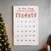 A personalized Christmas countdown calendar hanging on a gray wall next to a decorated Christmas tree. The white fabric calendar features the text 'The Nolan Family Christmas Countdown' in red and brown, a row of five cartoon reindeer, and numbered pockets from 1 to 25.