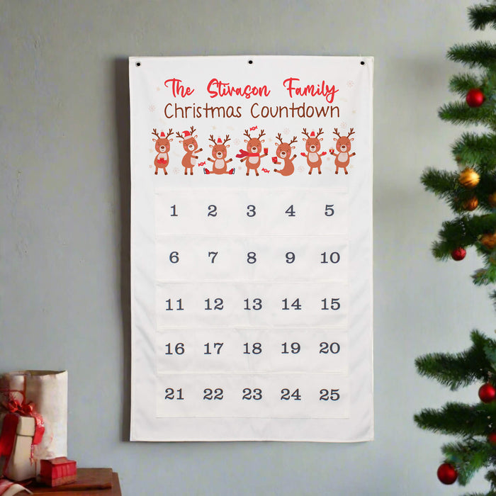 A white fabric Christmas countdown calendar hanging on a light gray wall, with red text reading 'The Stivason Family Christmas Countdown,' five cartoon reindeer, and numbered days 1 through 25. Part of a decorated Christmas tree and wrapped gifts are visible at the edges.