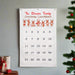 A white fabric Christmas countdown calendar hanging on a light gray wall, with red text reading 'The Stivason Family Christmas Countdown,' five cartoon reindeer, and numbered days 1 through 25. Part of a decorated Christmas tree and wrapped gifts are visible at the edges.